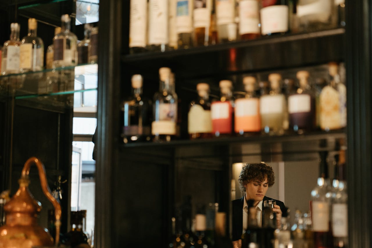 our-story A man with curly hair enjoys a drink at an elegant bar surrounded by assorted bottles.